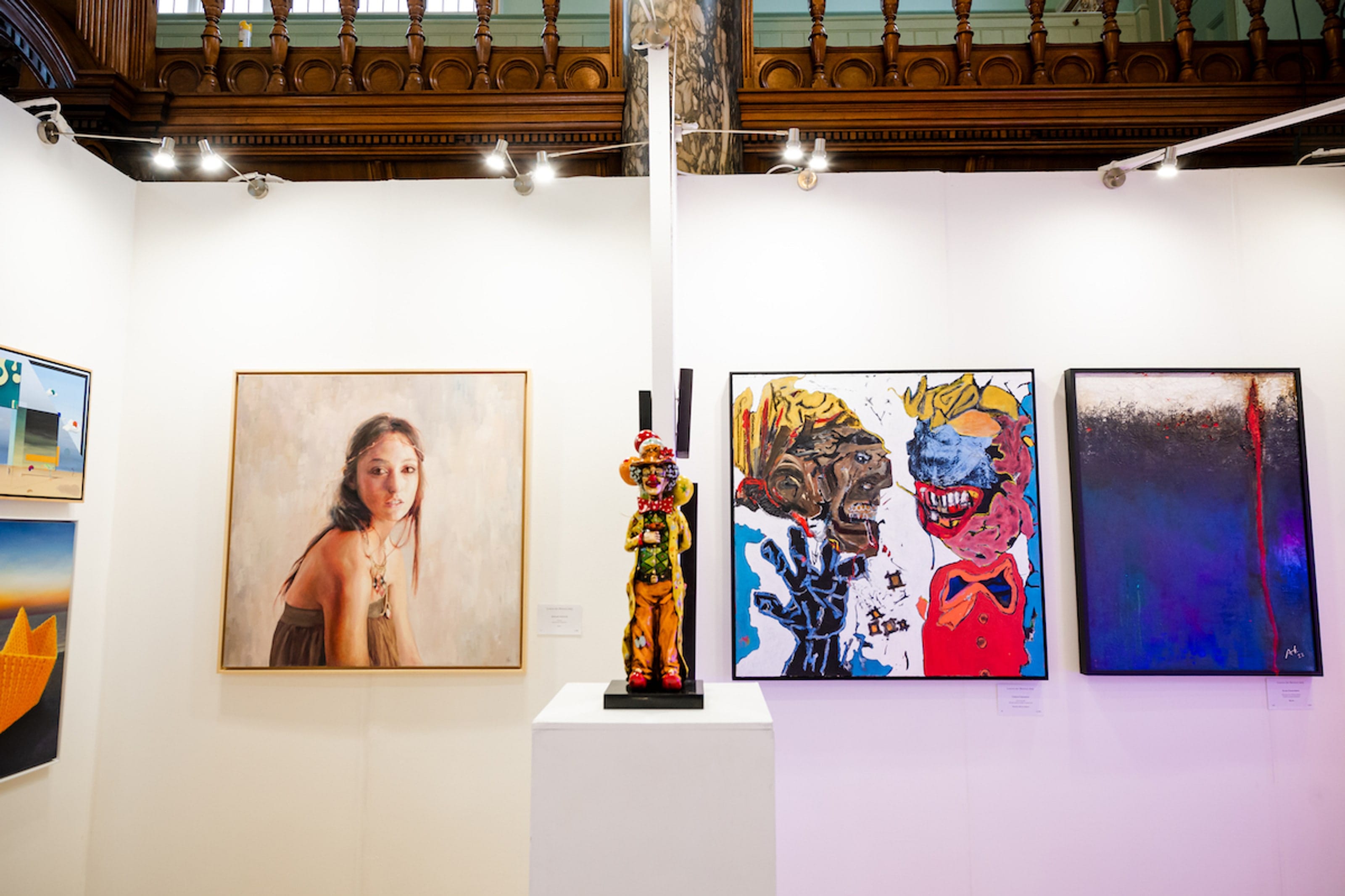 A booth hung with a portrait, two bright figurative canvases and a deep blue abstract, a painted figure sculpture between them.