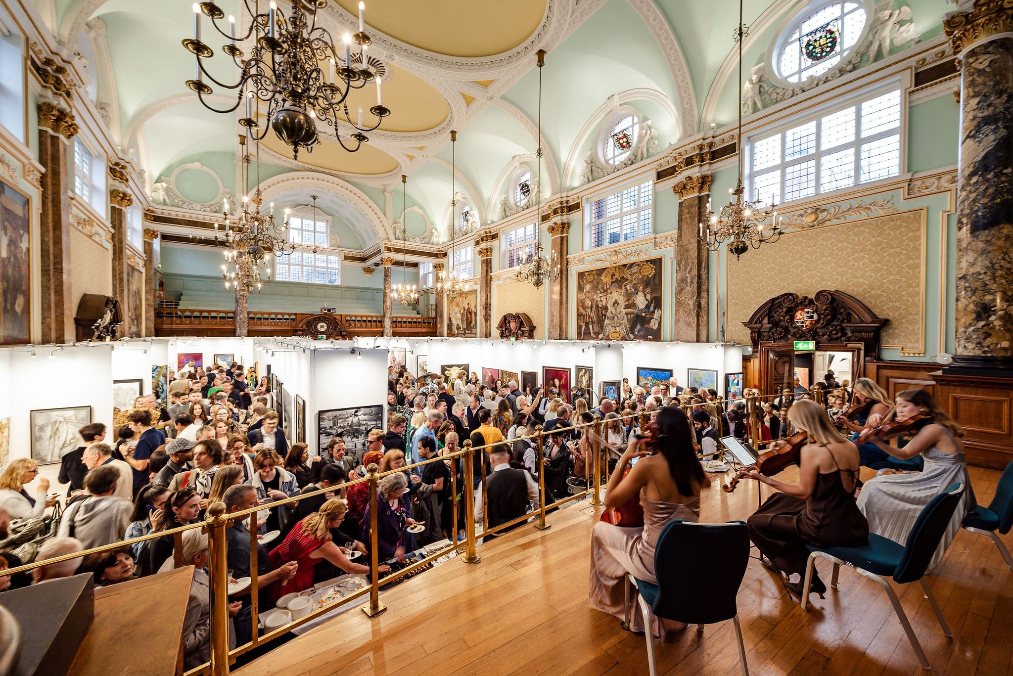 The main hall packed for the Biennale's prizegiving, seen past a string quartet playing at the balcony rail, with the hang, the chandeliers and the gilded vault beyond.