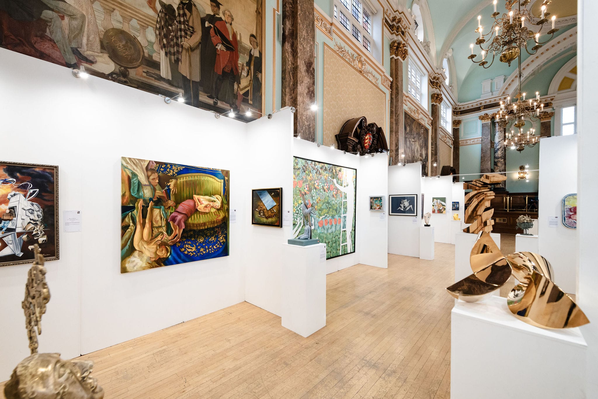 The 2025 hang along the length of the hall: paintings on white walls, gilded bronze sculpture in the foreground and the chandeliers beyond.