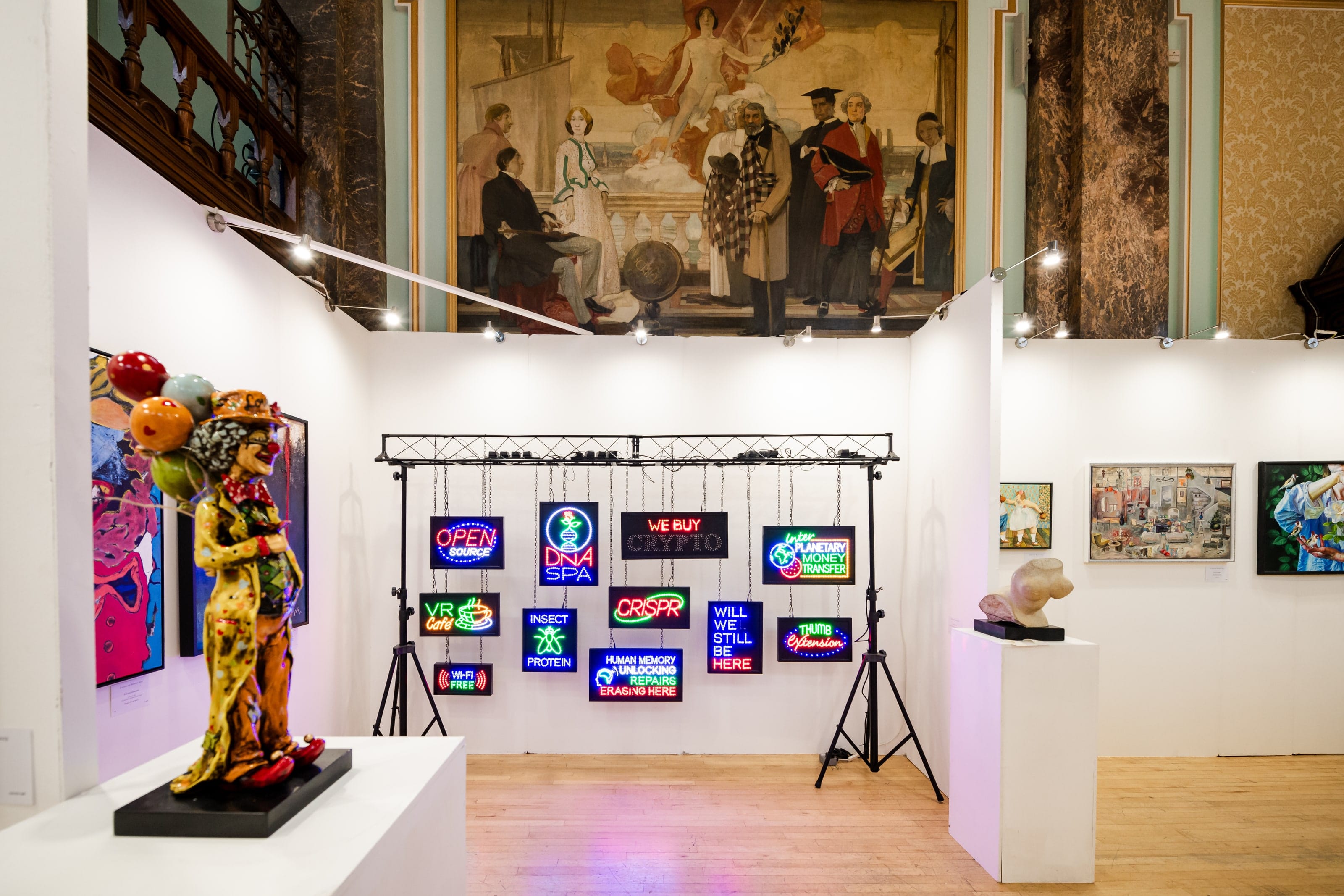 A booth of neon text works on a lighting rig, a figurative sculpture on a plinth beside it and a painted mural above the walls.