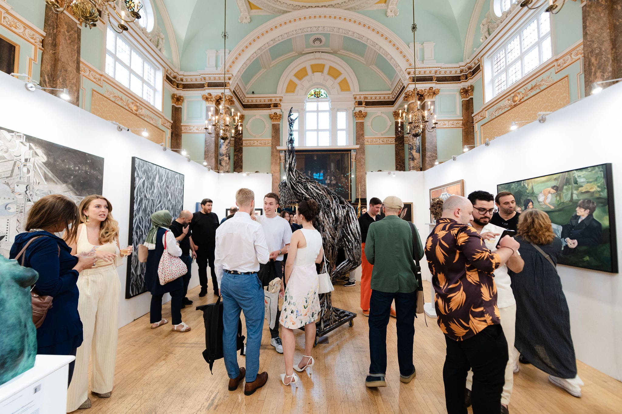 Visitors filling the main hall at the private view, the hang around them and the painted and gilded vault above.