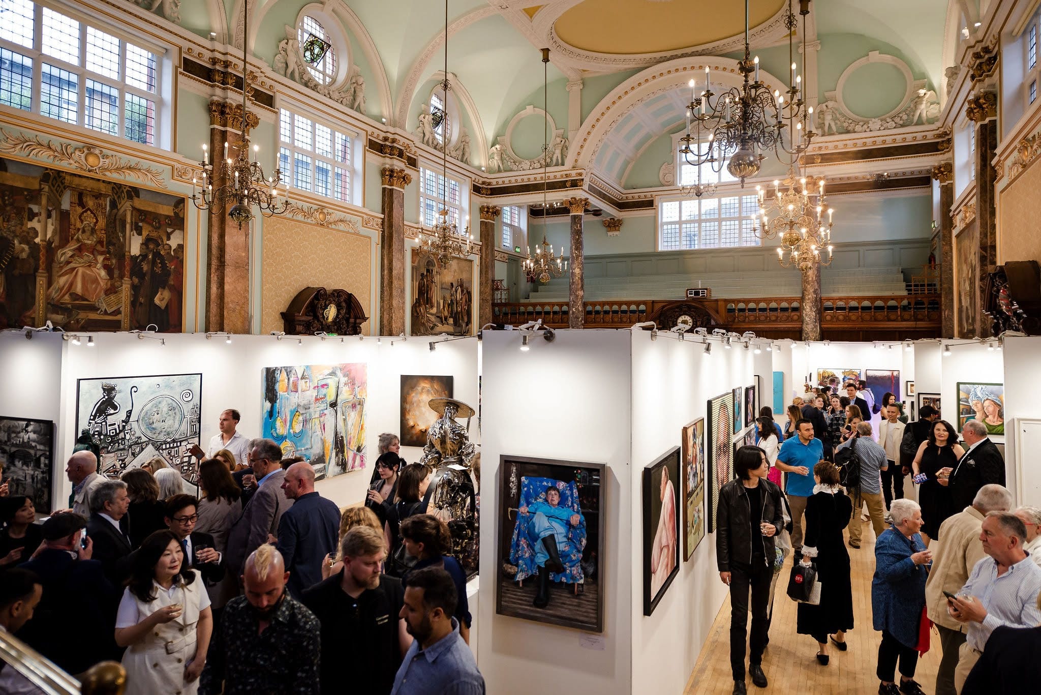 The main hall full for an opening, seen from the side balcony: visitors moving between the white walls of hung work beneath chandeliers and painted murals.