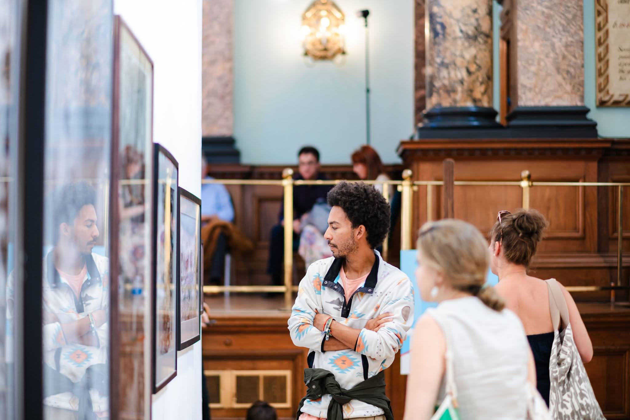 A visitor standing in front of a hung painting, considering it, the panelled hall and other visitors behind.