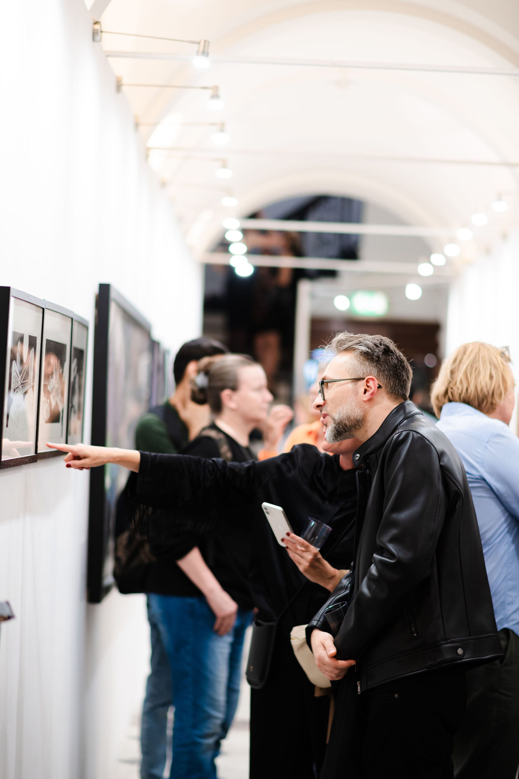A visitor pointing out a detail in a row of framed photographs, under the lights of the vaulted corridor.