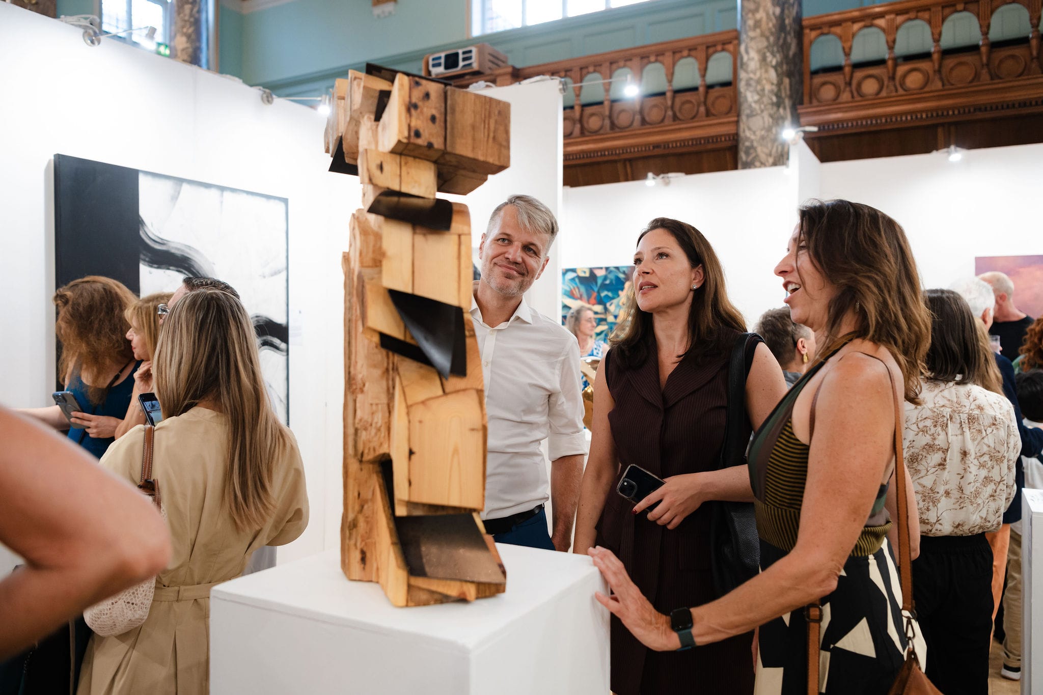 Visitors talking and laughing around a carved wooden sculpture at the private view, phones in hand.